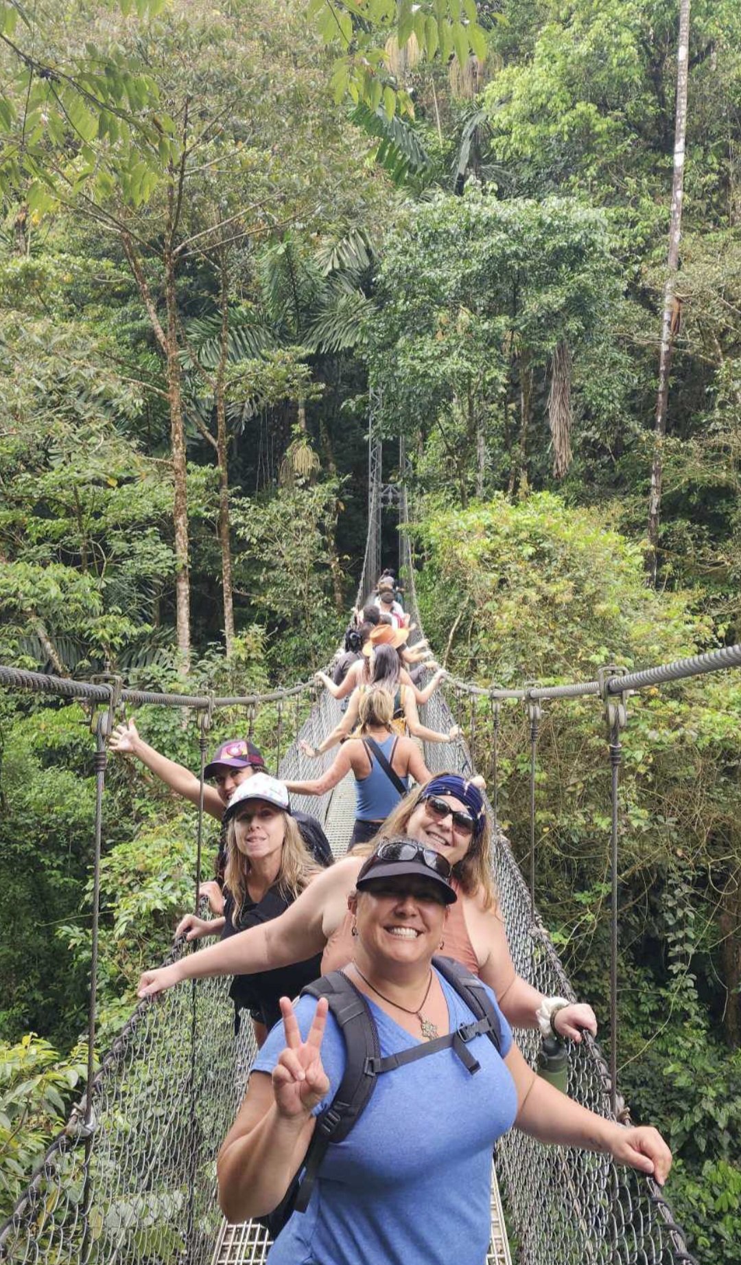 Group of tourists on a suspension bridge in a lush rainforest, smiling and making peace signs.