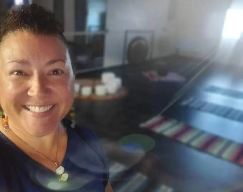 A smiling woman with short dark hair and colorful earrings standing in a room with a glass display case and a colorful striped rug.