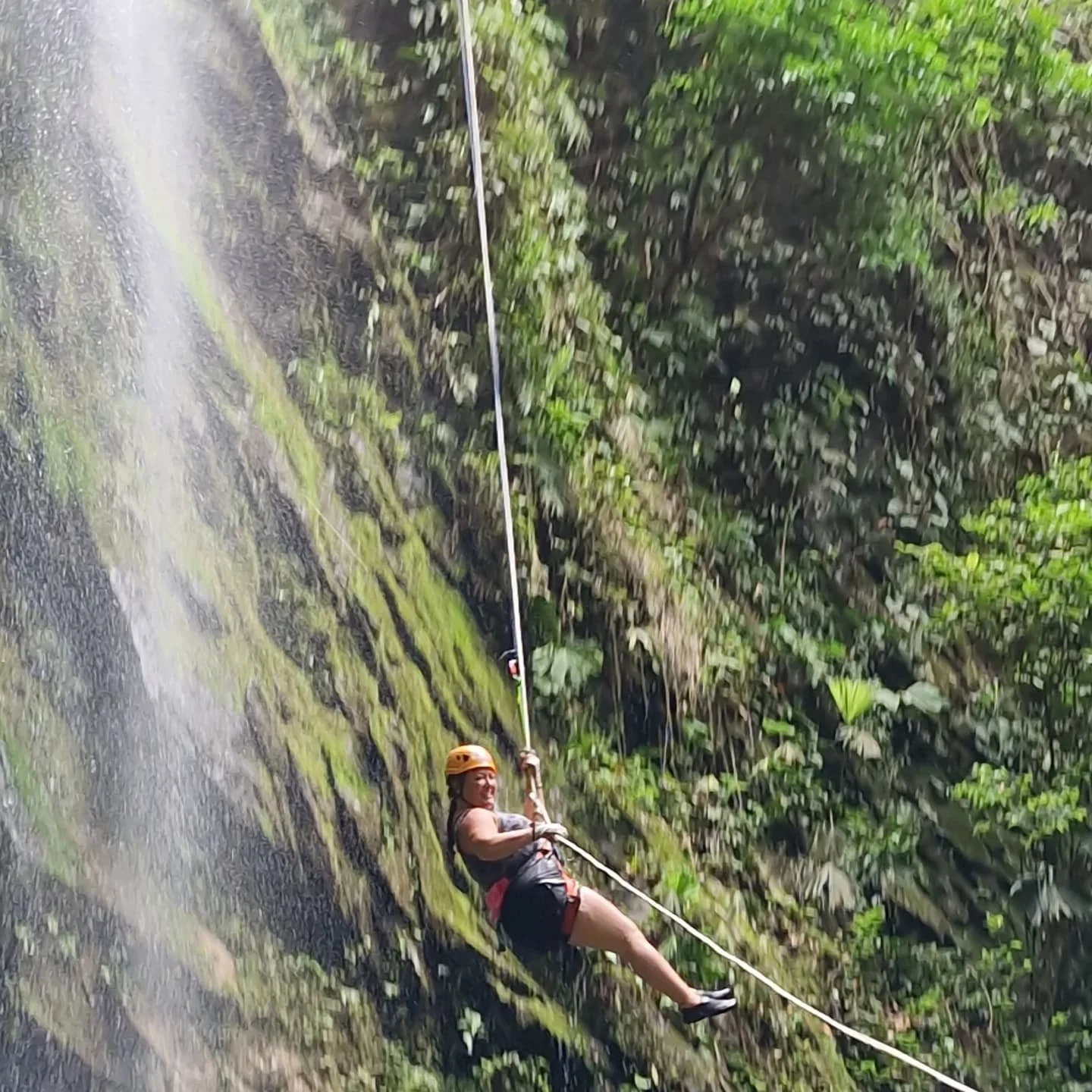 Person wearing orange helmet and black attire enjoying zip lining across a lush green forest.