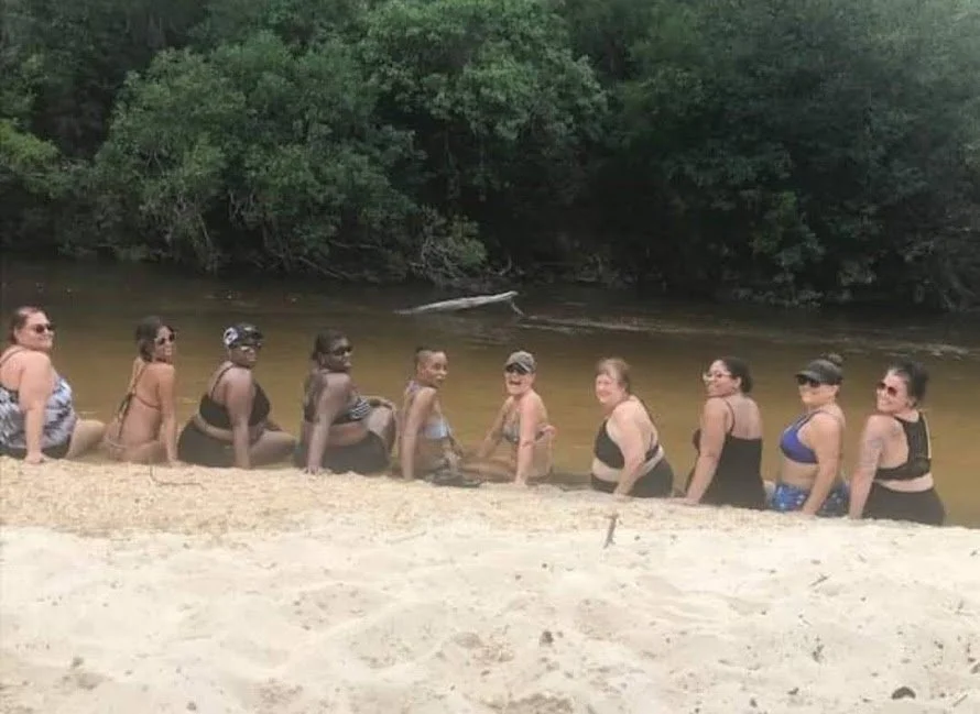 A group of women in swimsuits standing in a line in a river near a sandy beach with green trees in the background.