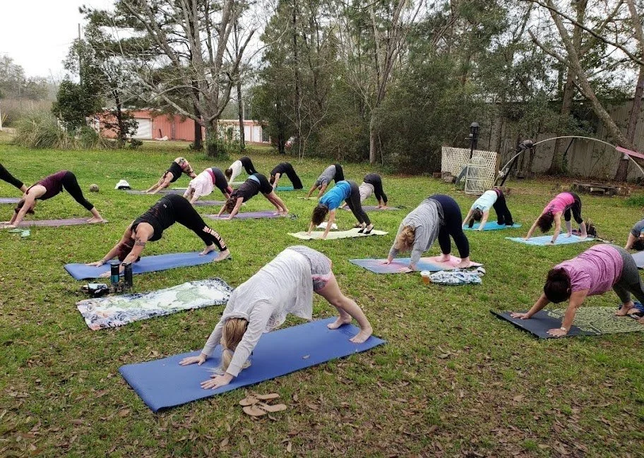 Group of women doing outdoor yoga in a grass backyard with trees and a wooden fence.