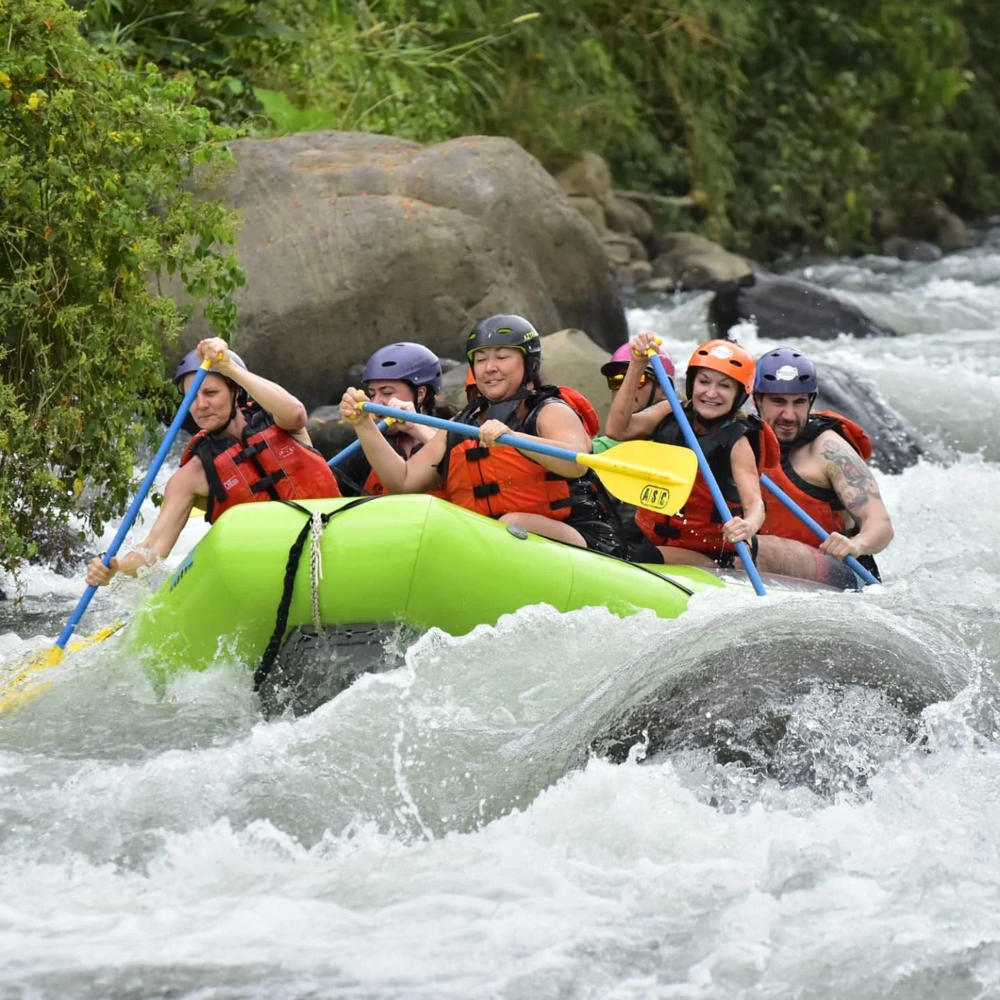 Group of six people white water rafting on a river, wearing life jackets and helmets, holding paddles, with trees and rocks in the background.