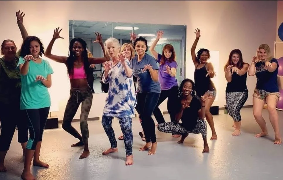 A group of women and girls in a dance or yoga class, posing enthusiastically in a studio with a mirror behind them.