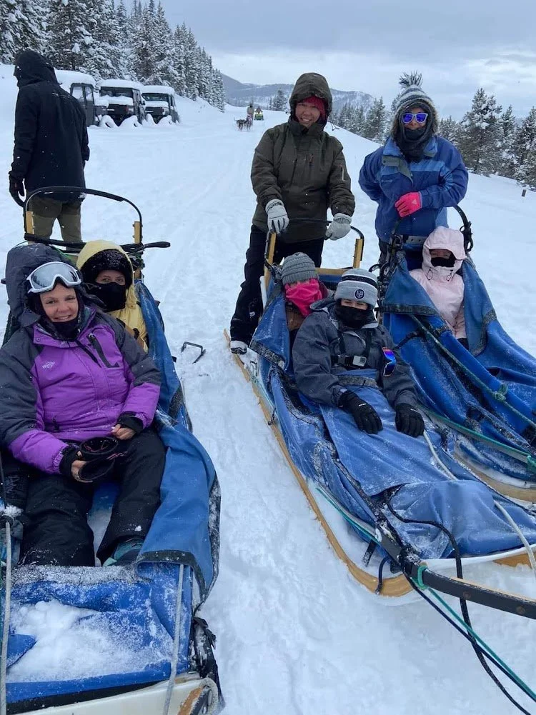 Group of people in winter gear with sleds on snow-covered trail, snowy trees, and mountains in the background.