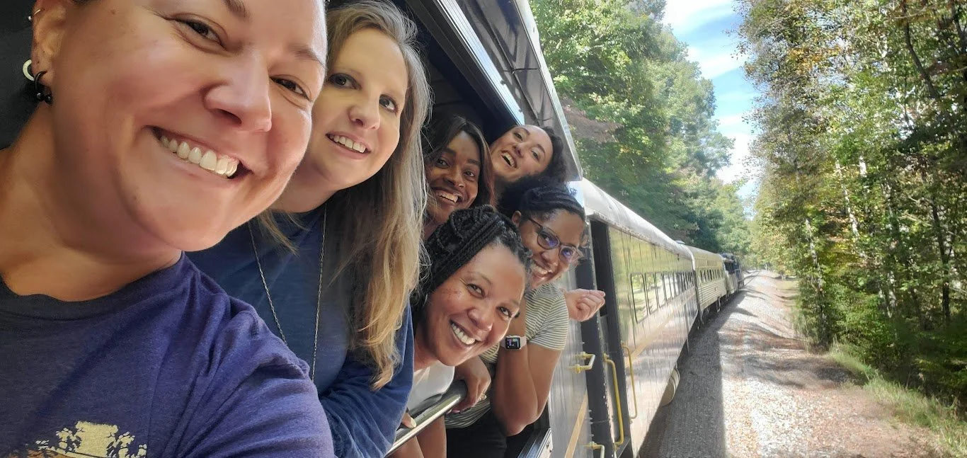 Group of people smiling and looking out a train window as the train travels through a wooded area.