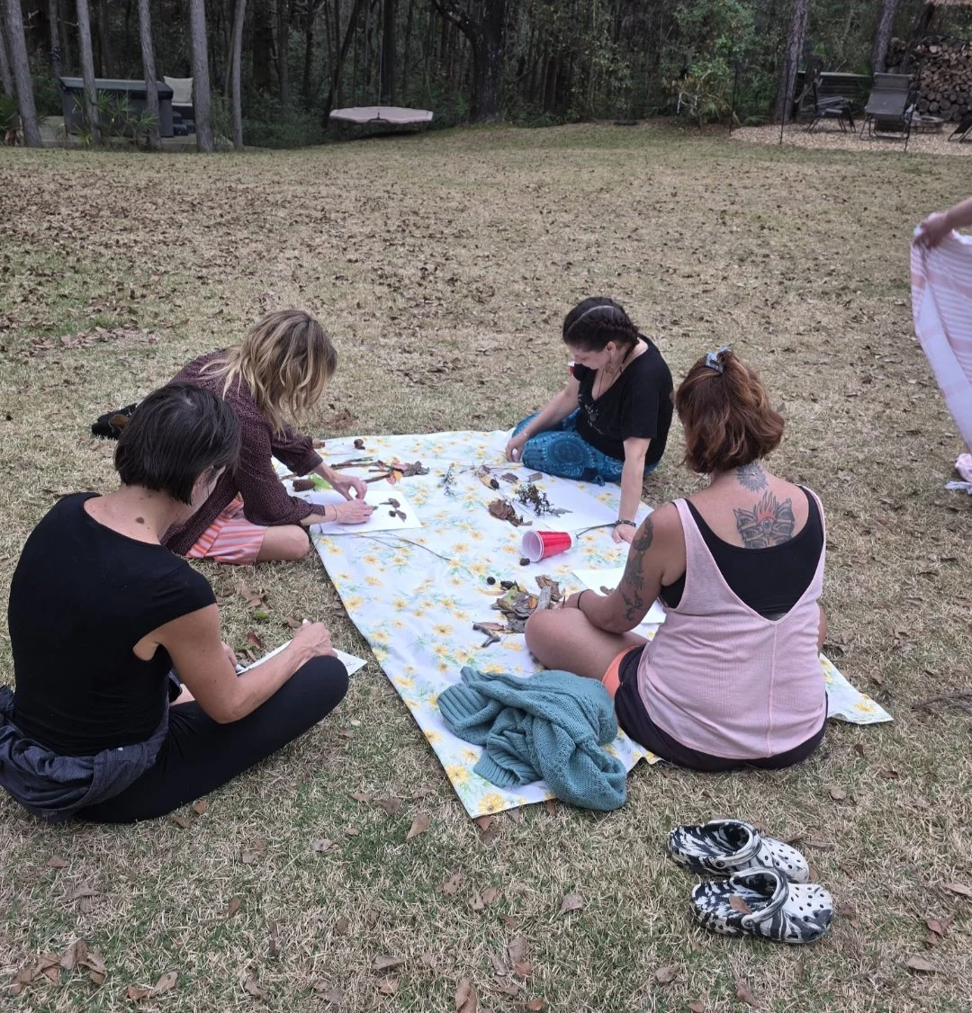 Four women sit on a blanket outdoors in a backyard, drawing or working on art projects with leaves and natural materials, with a forested area in the background.