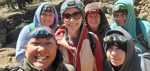 Group of six people outdoors with hiking gear, smiling and wearing headlamps.