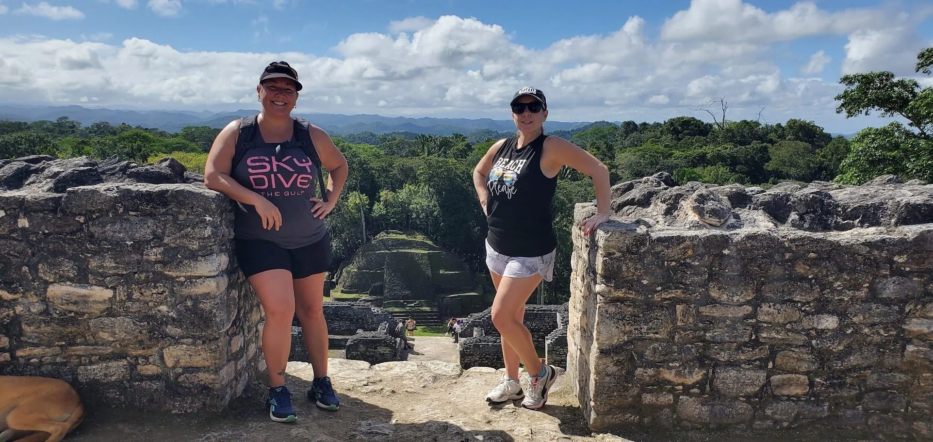 Two women standing between stone ruins at an ancient archaeological site with lush green jungle and mountains in the background under a cloudy sky.