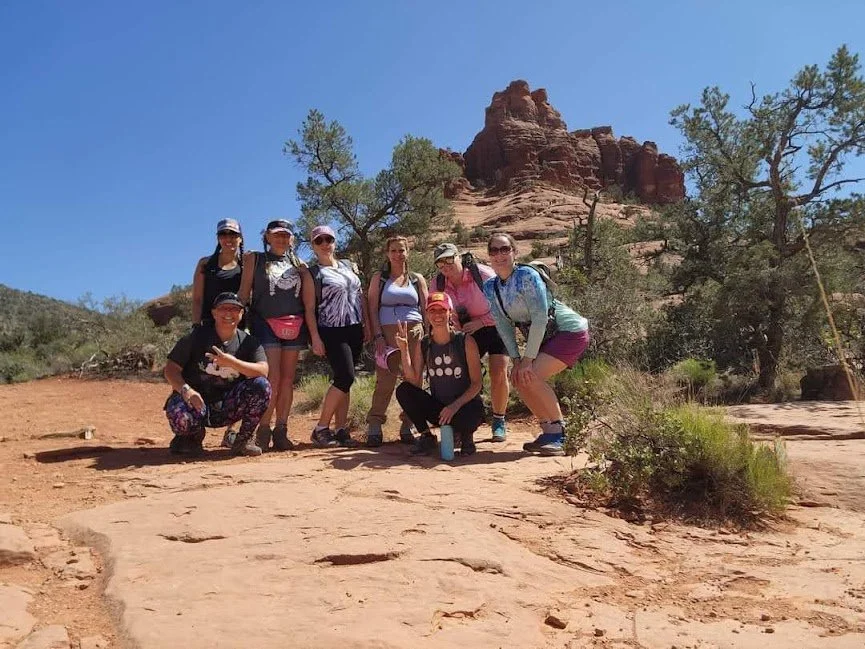 Group of eight women on a desert hike with red rock formations and trees in the background.