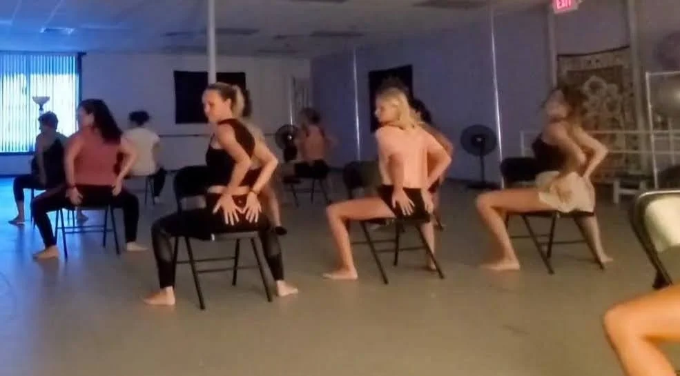 Group of women practicing chair dance in a dance studio with mirrors and blue lighting.