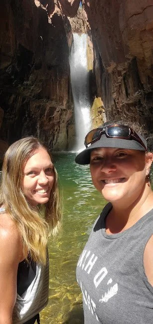 Two women taking a selfie in front of a waterfall inside a canyon, with water at their feet and rocky walls.