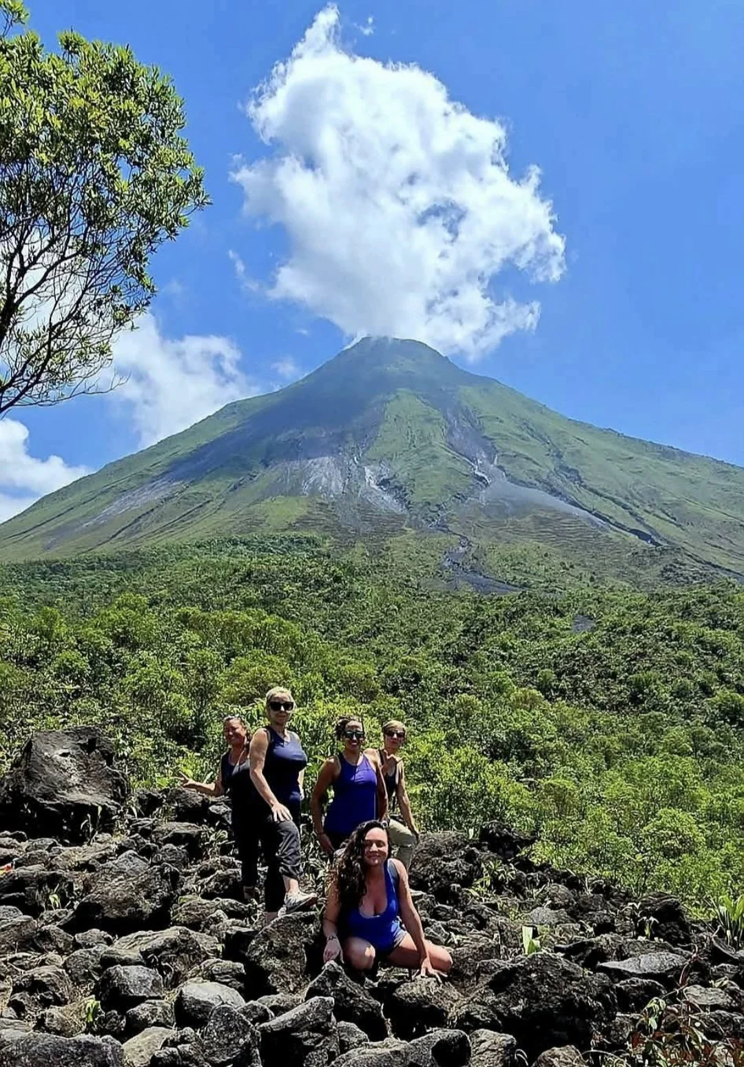 A group of five women hiking on rocky terrain with a lush green volcanic mountain in the background, topped with a small cloud against a blue sky.