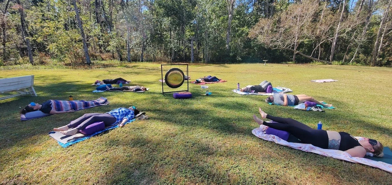 Group of people lying on yoga mats outdoors in a grassy field, practicing a relaxation or meditation session in front of a gong.