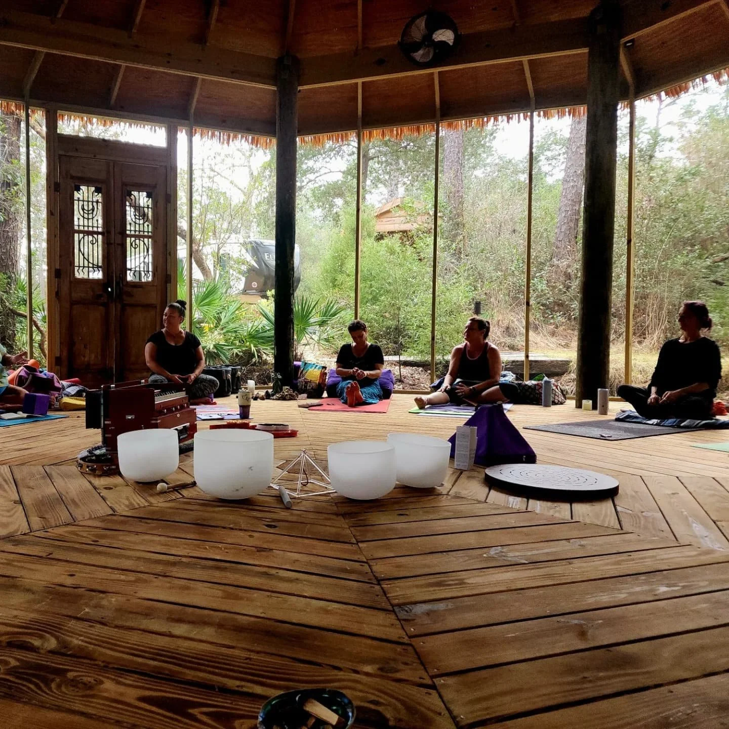 A group of five people sits cross-legged on yoga mats inside a wooden open-air structure surrounded by trees. Musical instruments and singing bowls are on the floor in the foreground.