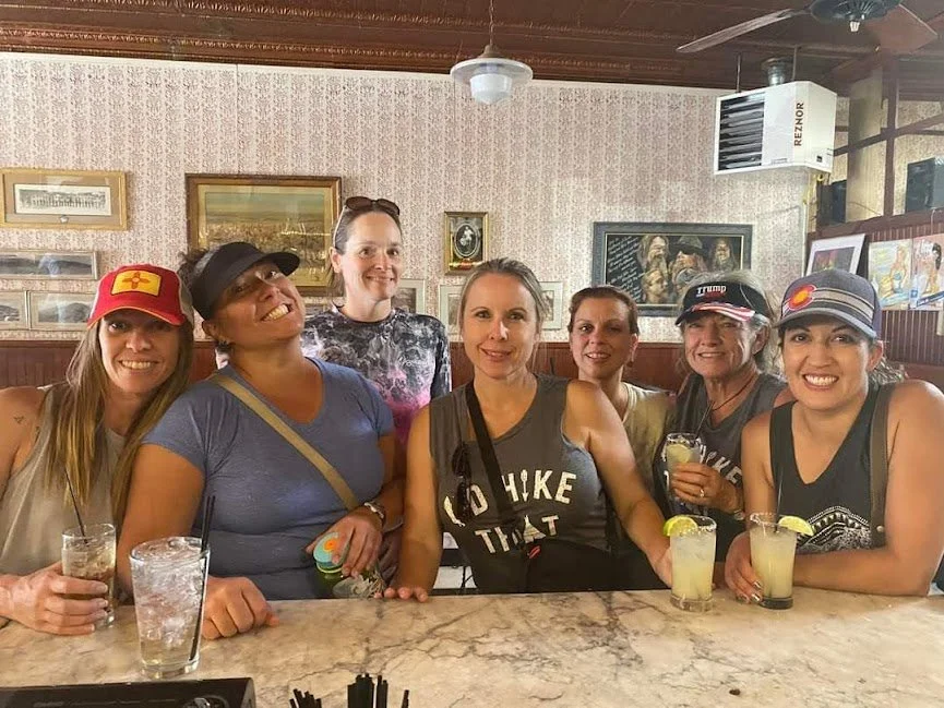 Six women sitting at a bar, smiling, with drinks in their hands, in a cozy restaurant with framed pictures on the wall.