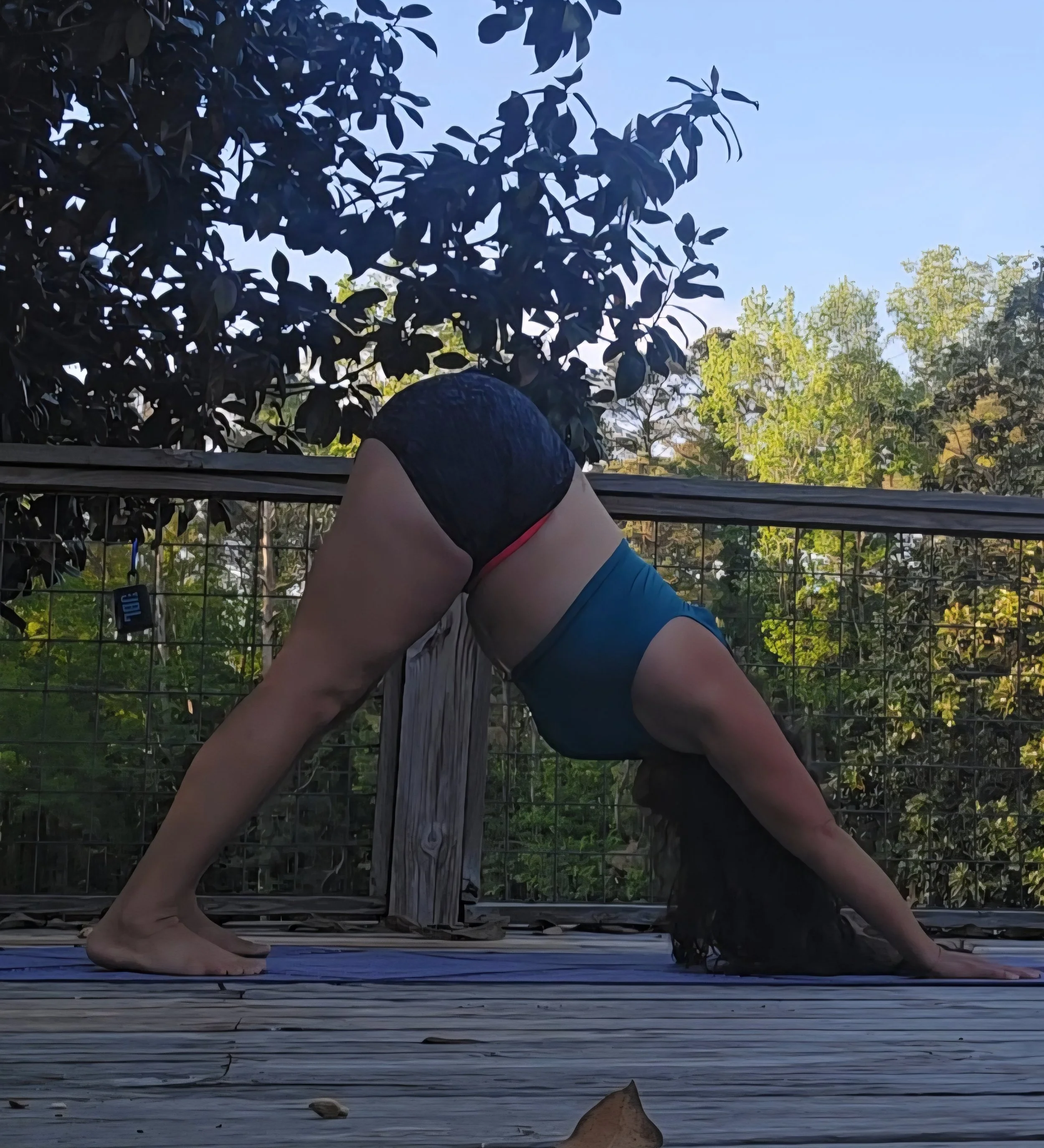View of a person doing yoga or stretching on a wooden deck outdoors, with trees and a clear blue sky in the background.