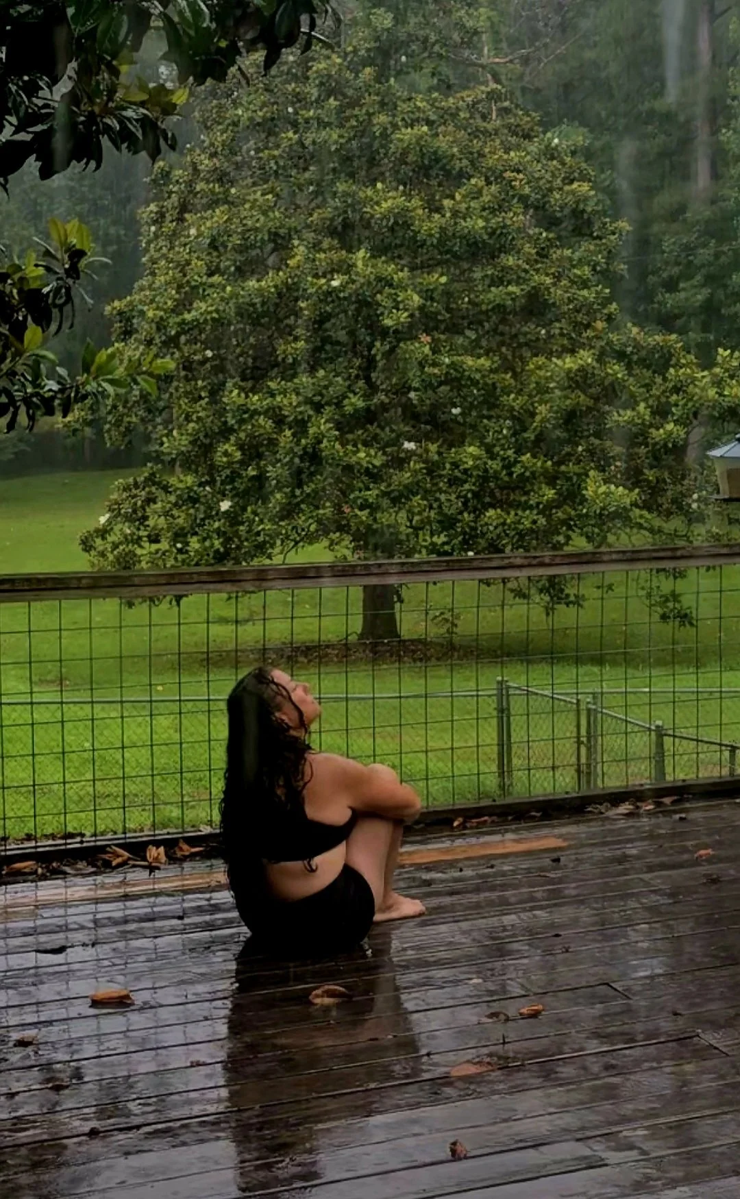 A woman with wet hair and black clothing sitting on a wooden deck, looking up, with a green tree and grass in the background, and rain falling around her.