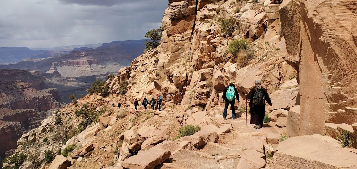 Hikers walking along a rocky trail on the side of a canyon with layered rock formations and sparse vegetation.