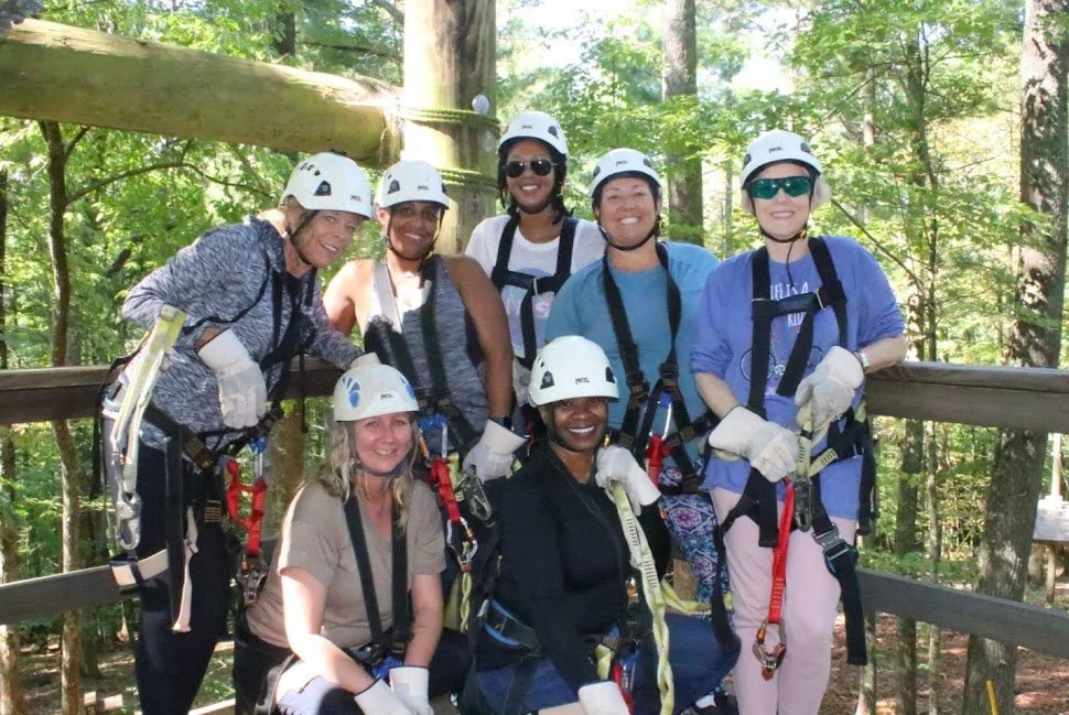 Group of women in safety helmets and harnesses standing on a wooden platform in a forest.