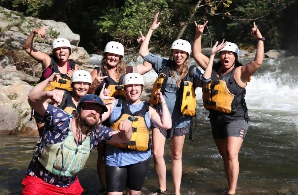 Group of six people wearing helmets and life jackets on a river, posing with raised arms and smiling.