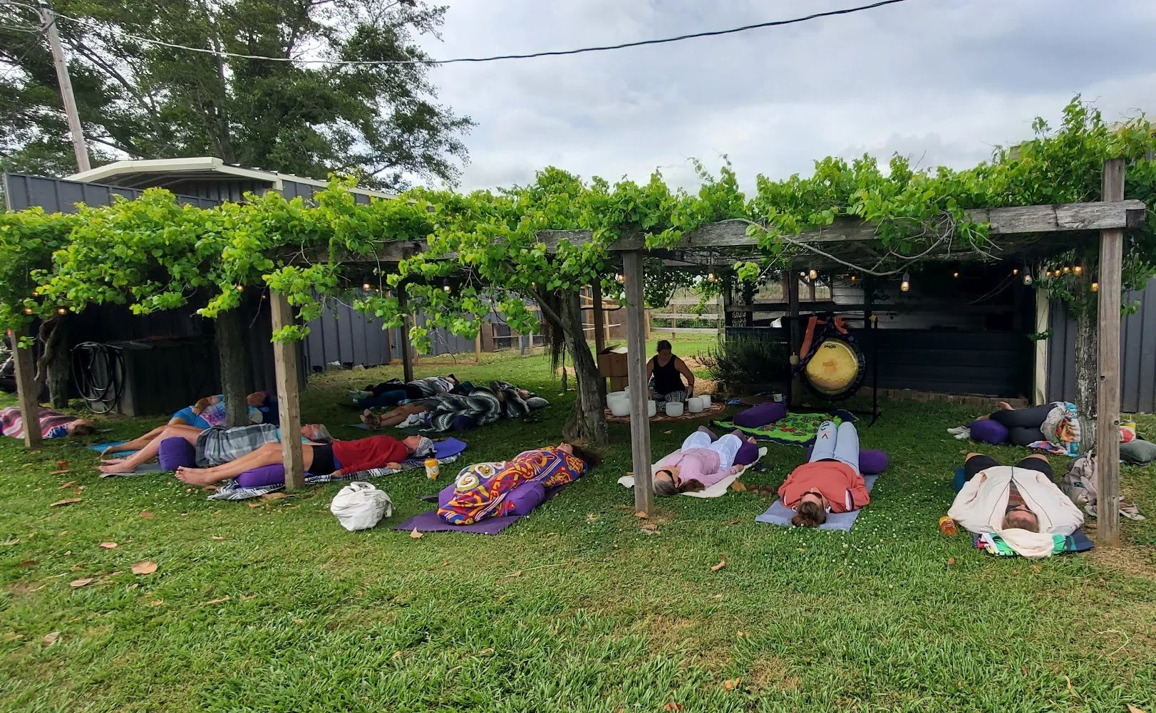 People participating in a group outdoor yoga or meditation session beneath a grapevine-covered wooden pergola on a grassy area, with cloudy sky overhead.