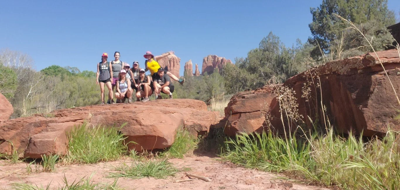 Group of nine people, mostly women, posing on large red rocks outdoors in a desert-like area with some trees and rocky formations in the background under a clear blue sky.
