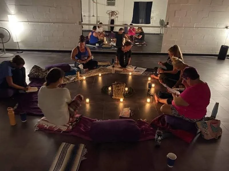 Group of women sitting in a circle on cushions with candles and books in a dimly lit studio, practicing meditation or yoga.