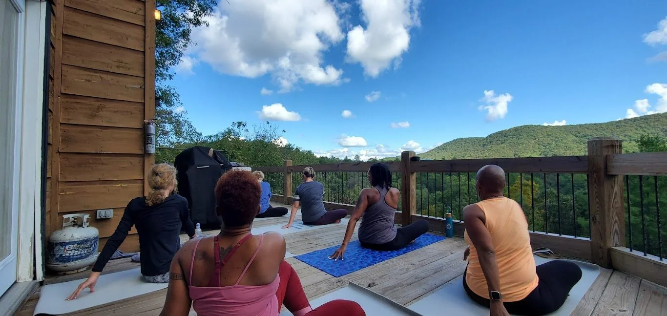 Six people practicing yoga on a wooden deck overlooking lush green mountains on a sunny day.