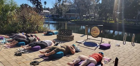 People practicing yoga outdoors on a paved area near a body of water, lying on yoga mats in a peaceful setting with trees and buildings in the background.