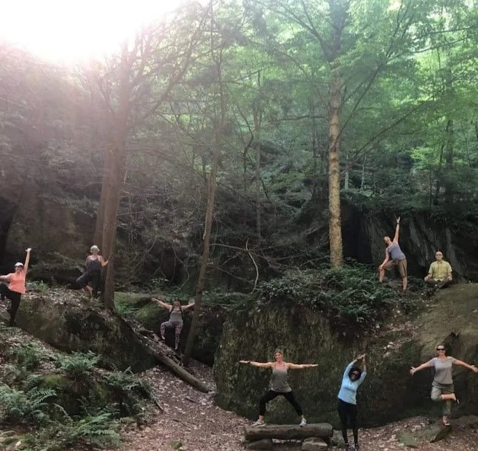 People practicing yoga outdoors in a lush, wooded area with large rocks and trees, with sunlight filtering through the leaves.