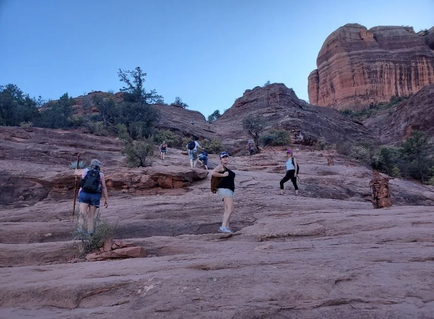 Group of hikers climbing a rocky slope in a desert landscape with large red rock formations and sparse vegetation, during daytime.