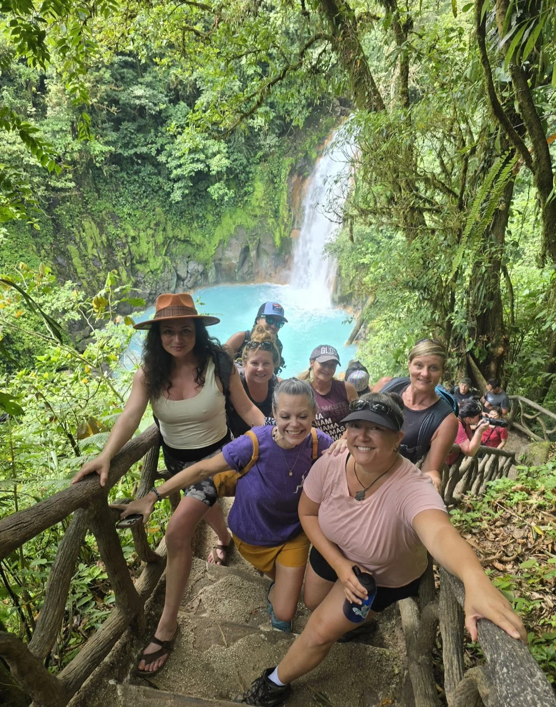 Group of people posing on a forest trail with a waterfall and blue pool in the background.