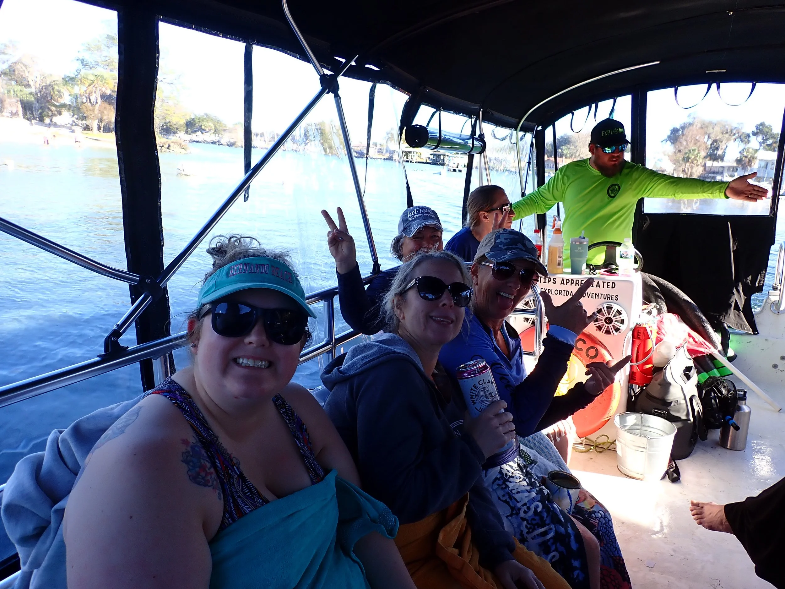Group of people on a boat taking a selfie. They are smiling and making peace signs. The background shows water, trees, and houses along the shoreline.