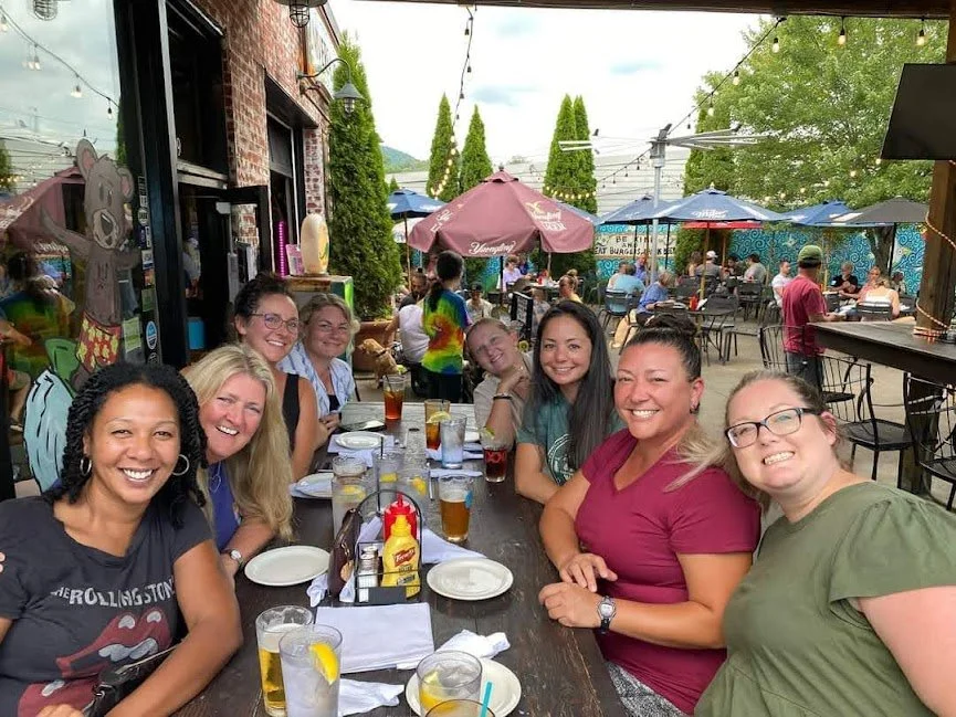 Group of nine women smiling at a long table outdoors at a restaurant or cafe with drinks, condiments, and plates, under string lights and umbrellas.