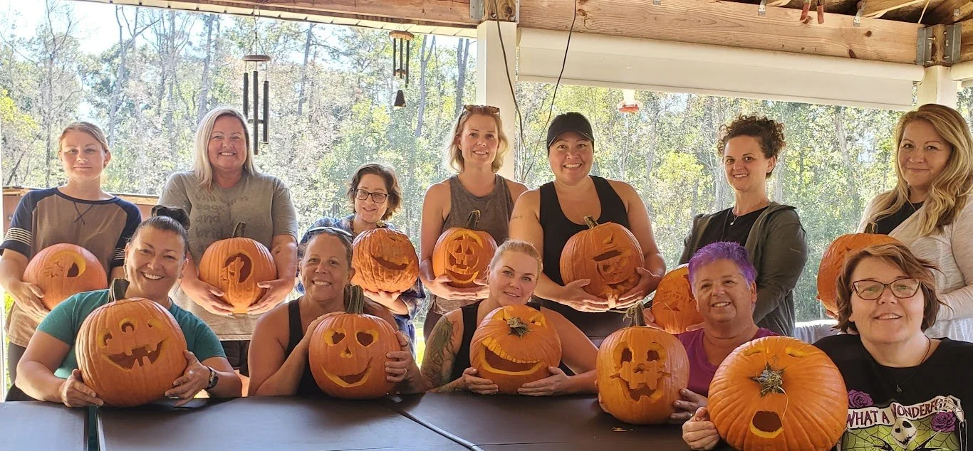 A group of women holding carved pumpkins with various faces, smiling for a photo indoors with windows revealing trees outside.