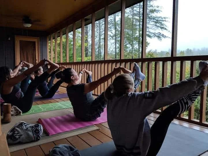 Group of women practicing yoga on mats inside a wooden room with large windows and a view of trees outside.