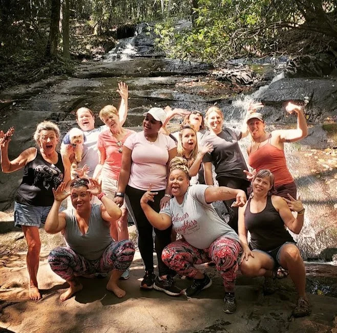 Group of women and children posing and smiling outdoors near a waterfall or stream in a forest setting.