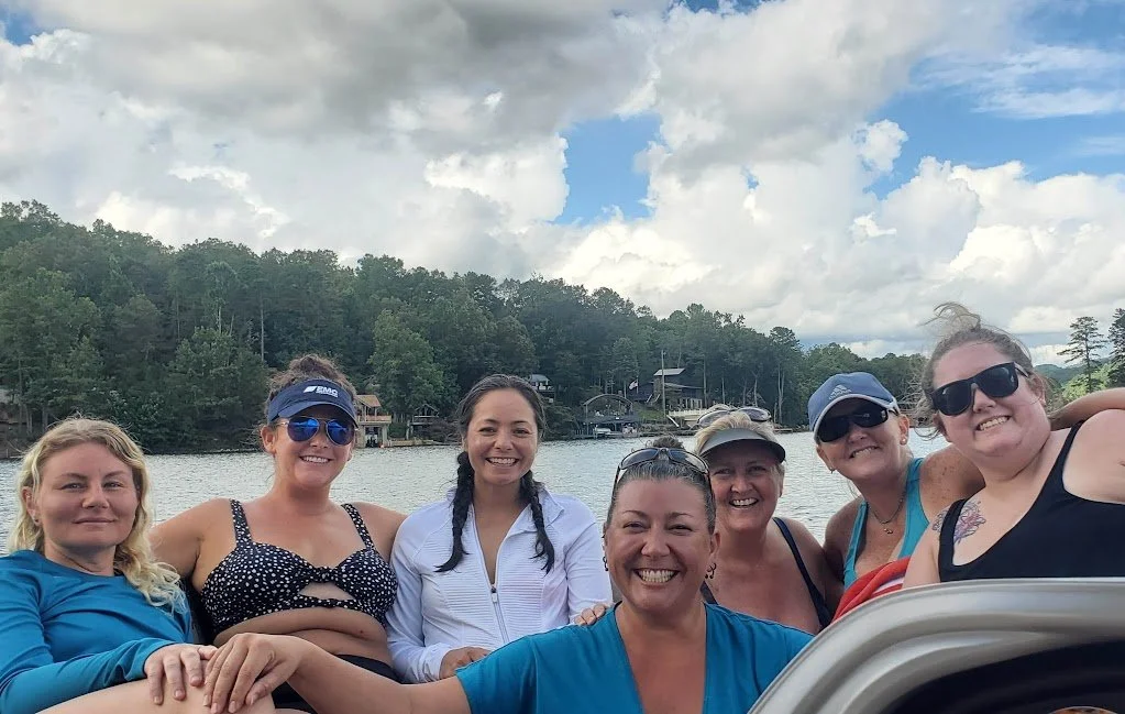 Group of women posing and smiling on a boat near a lakeside with trees and houses in the background under cloudy sky.