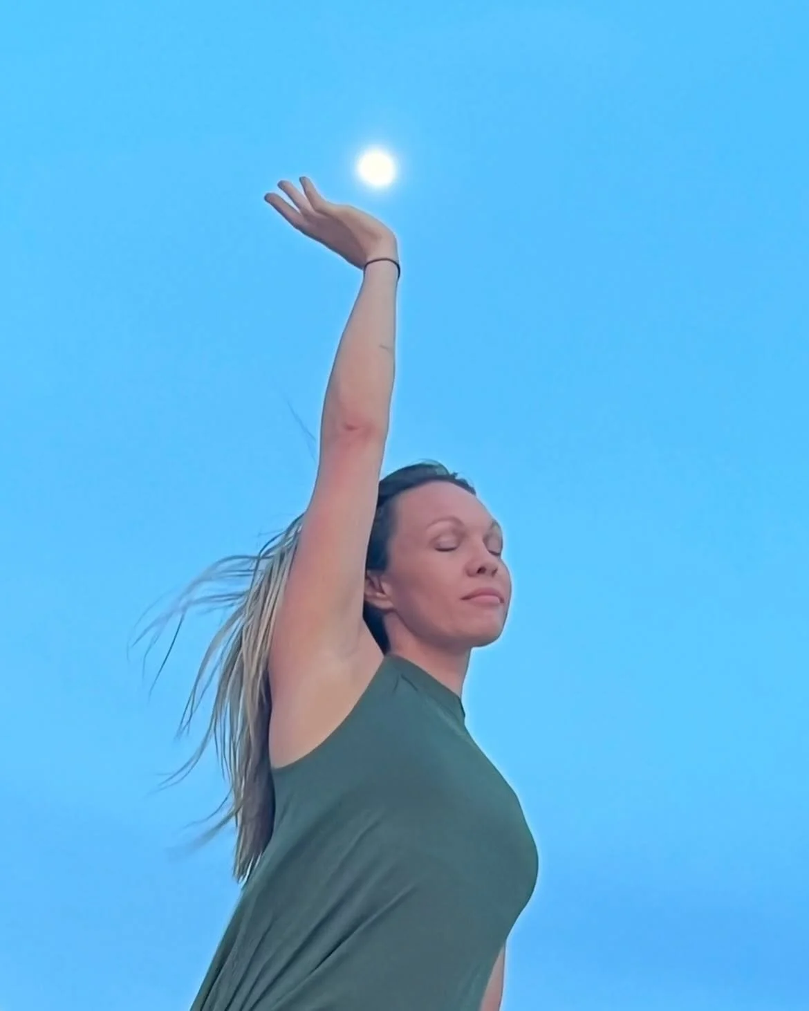 A woman practicing yoga outdoors during twilight, with her eyes closed and one arm raised, reaching towards the sky with the moon visible in the background.