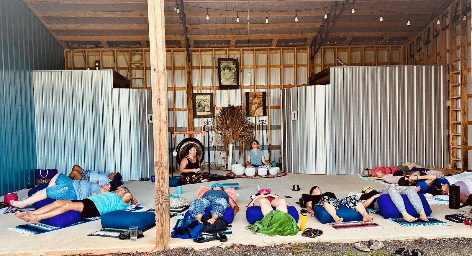 A group of people participating in a yoga class, lying on yoga mats and cushions inside a spacious barn with wooden and metal walls. Two instructors are at the front, one playing singing bowls and the other sitting on a cushion. The barn has a high ceiling with string lights and decorative pictures on the wall.