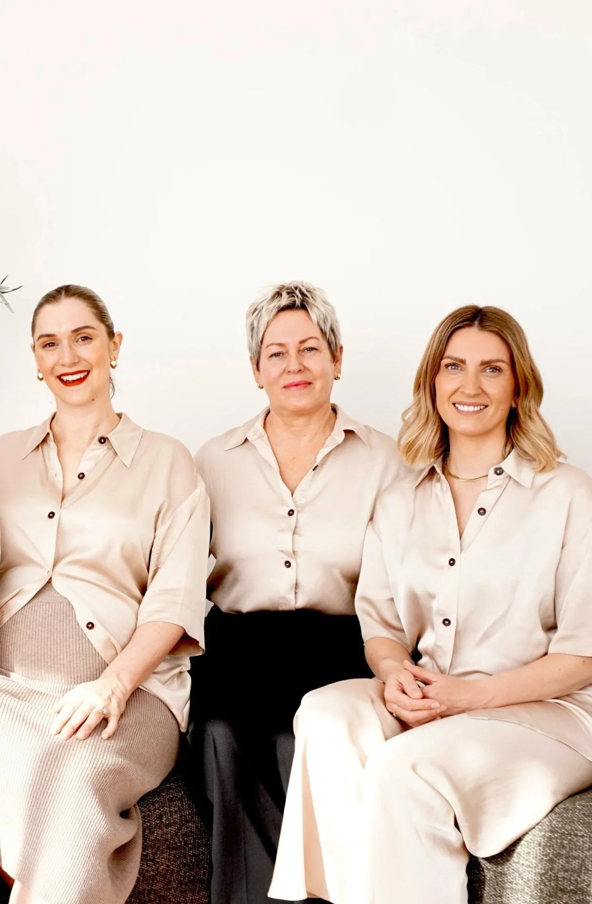Three women sitting together against a plain white background, all wearing light-colored, satin-like shirts and smiling.