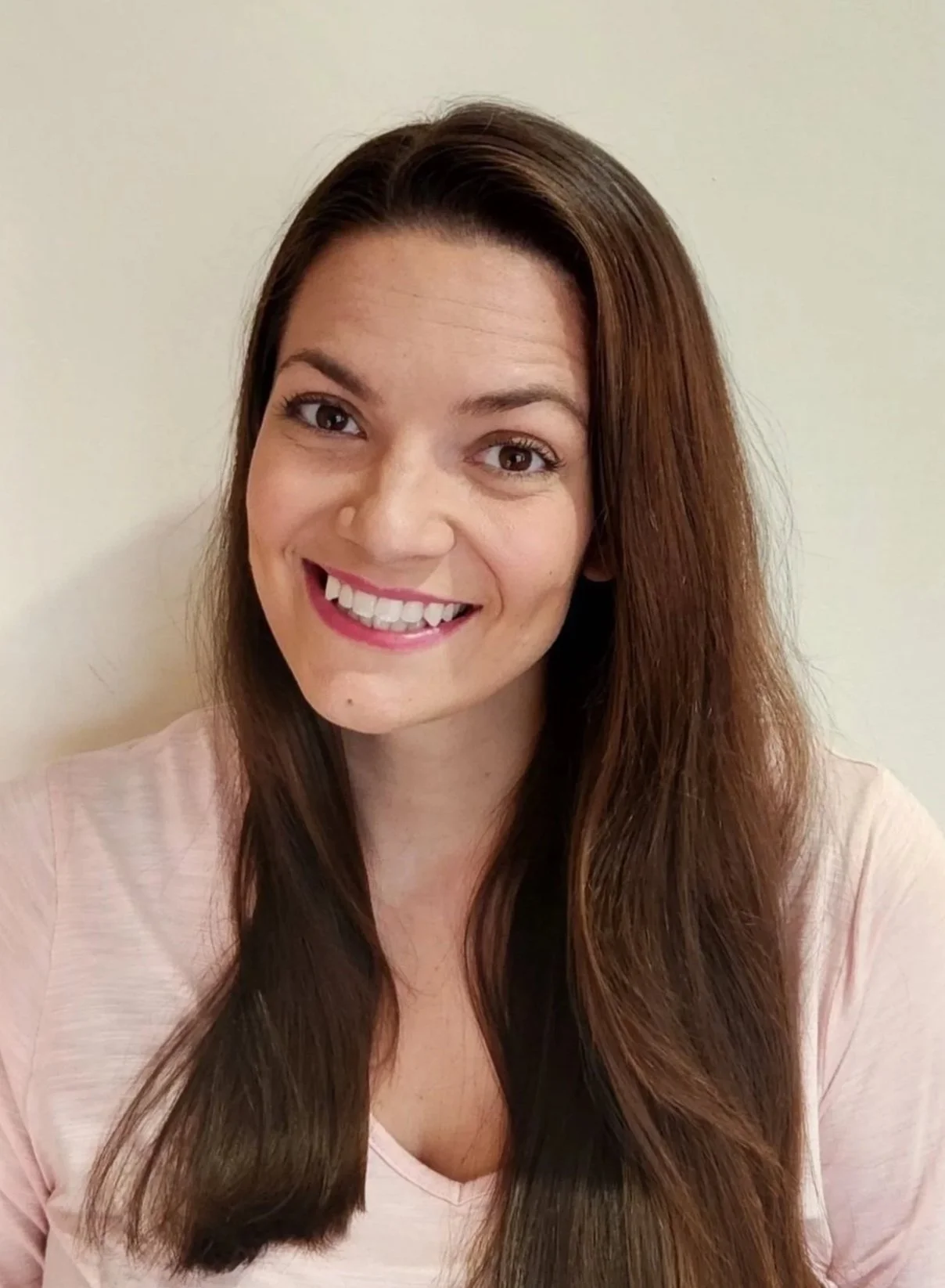 Portrait of a woman with long brown hair, smiling at the camera, wearing a light pink top, against a plain off-white background.