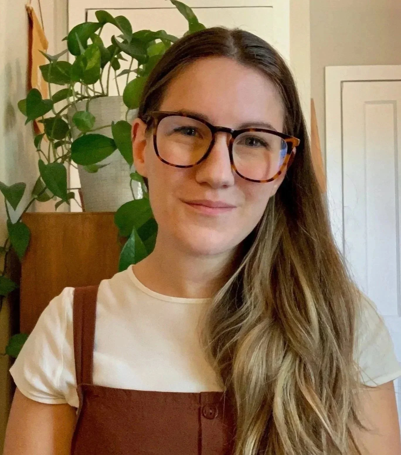 A woman with long, wavy brown hair, wearing black-rimmed glasses and a white t-shirt with brown overalls, standing indoors near a large green houseplant and a white door.