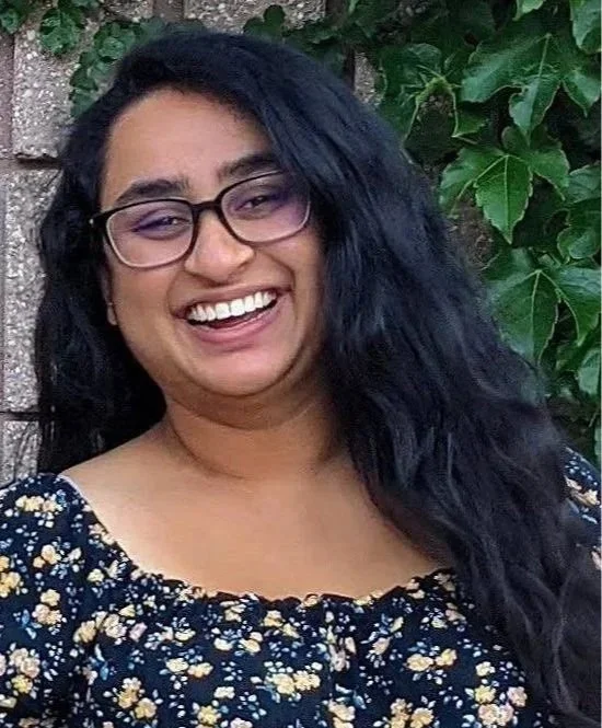 Smiling woman with glasses and long dark hair, wearing a floral dress, standing outdoors in front of green leaves and a brick wall.