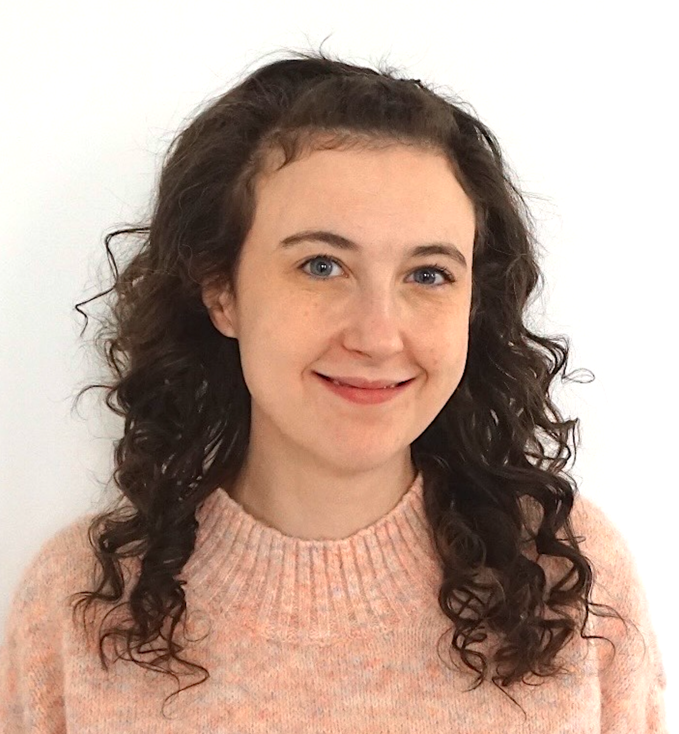 A woman with long curly brown hair, light skin, blue eyes, wearing a peach-colored sweater, smiling, standing against a plain white background.