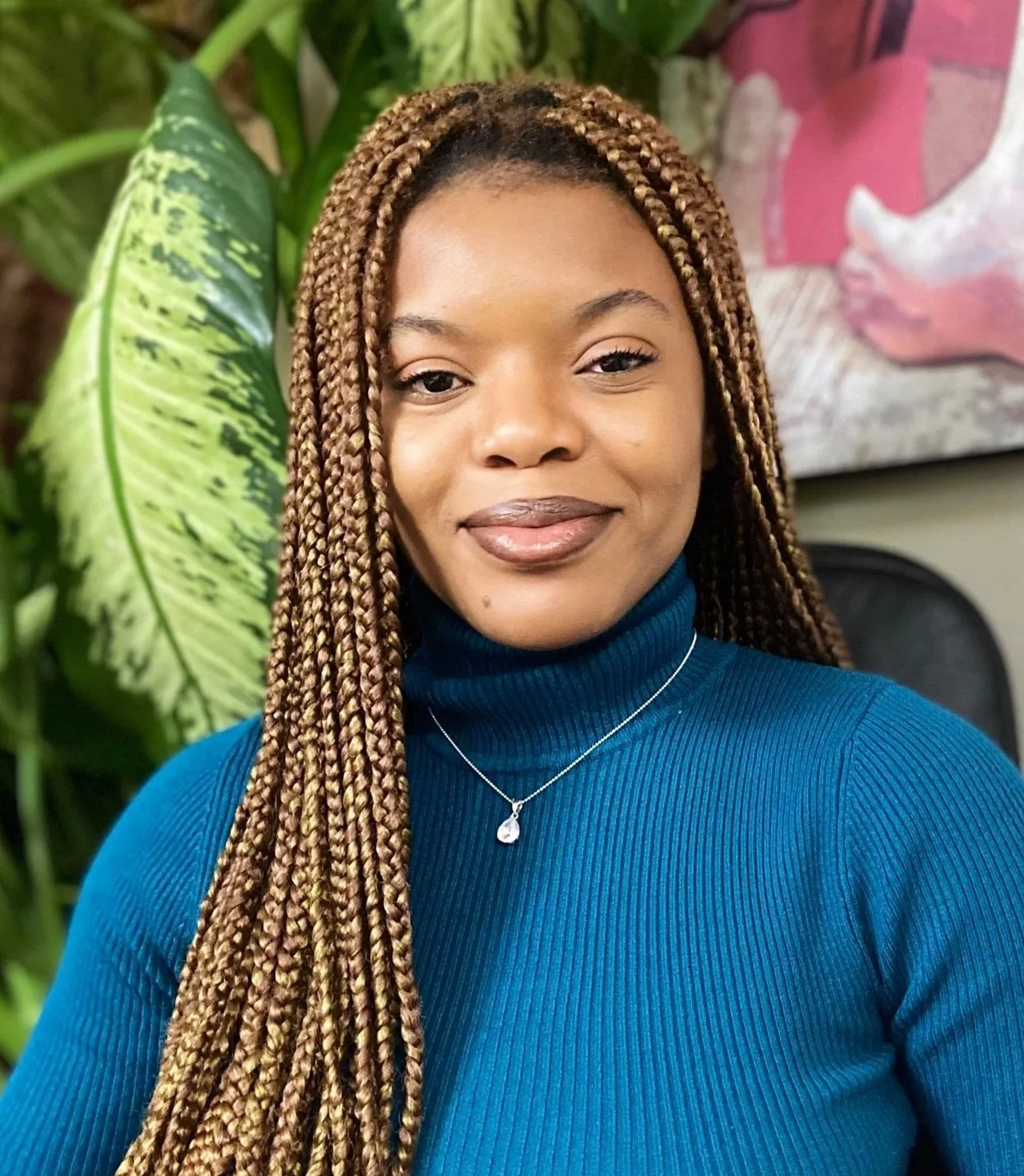 A young woman with long, braids, wearing a blue turtleneck sweater and a silver necklace with a teardrop pendant, smiling in front of green plants and a colorful painting.
