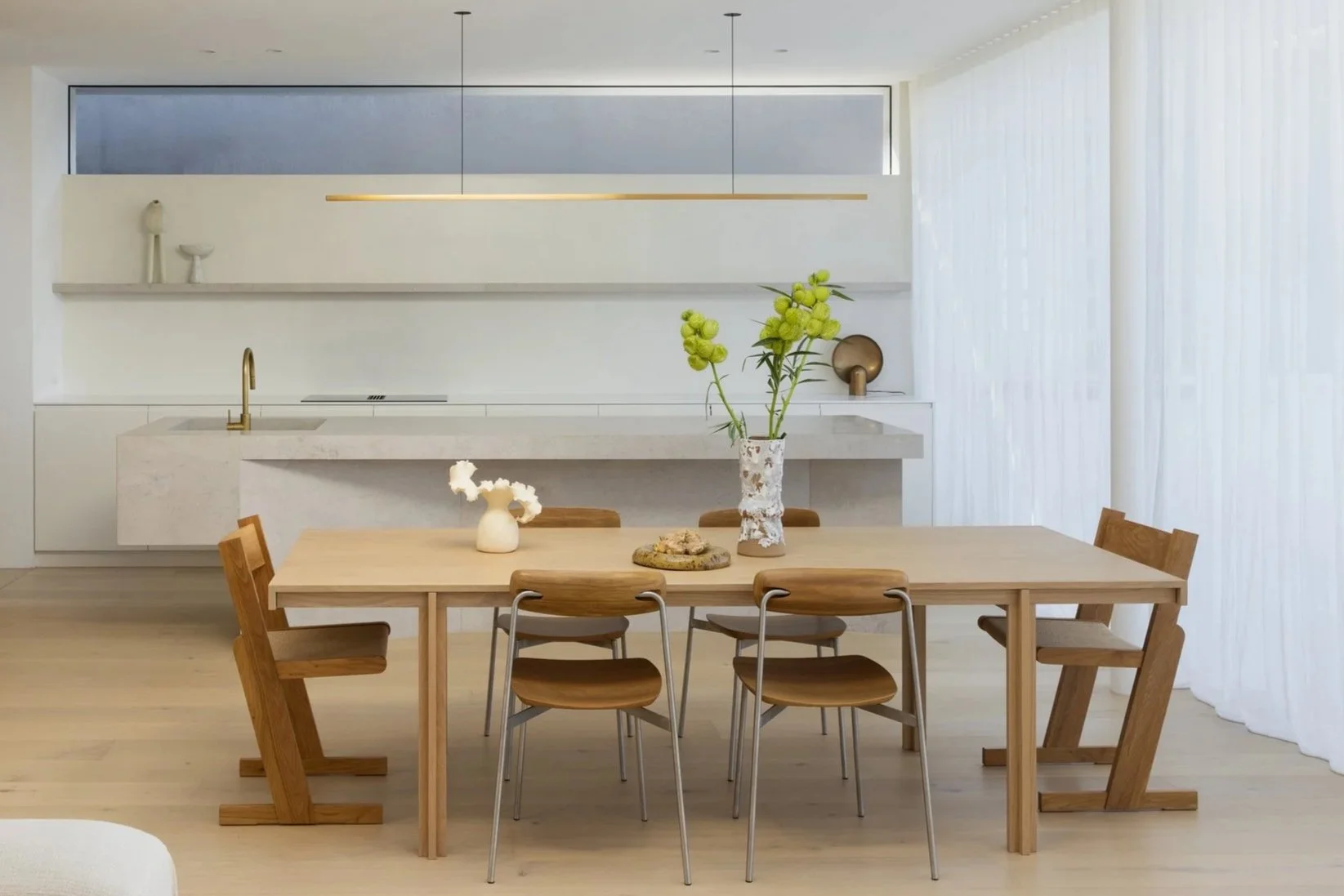 Modern minimalist kitchen and dining area with wooden table, six chairs, and decorative vases with flowers.