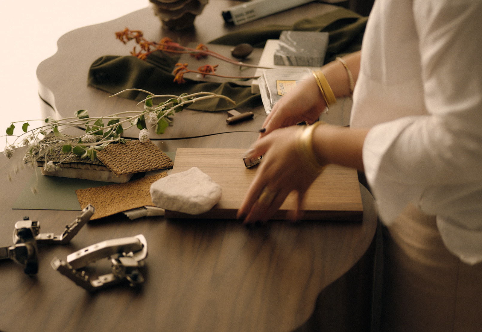 A person arranging various textured materials, plants, and stones on a wooden surface, with tools and supplies nearby.