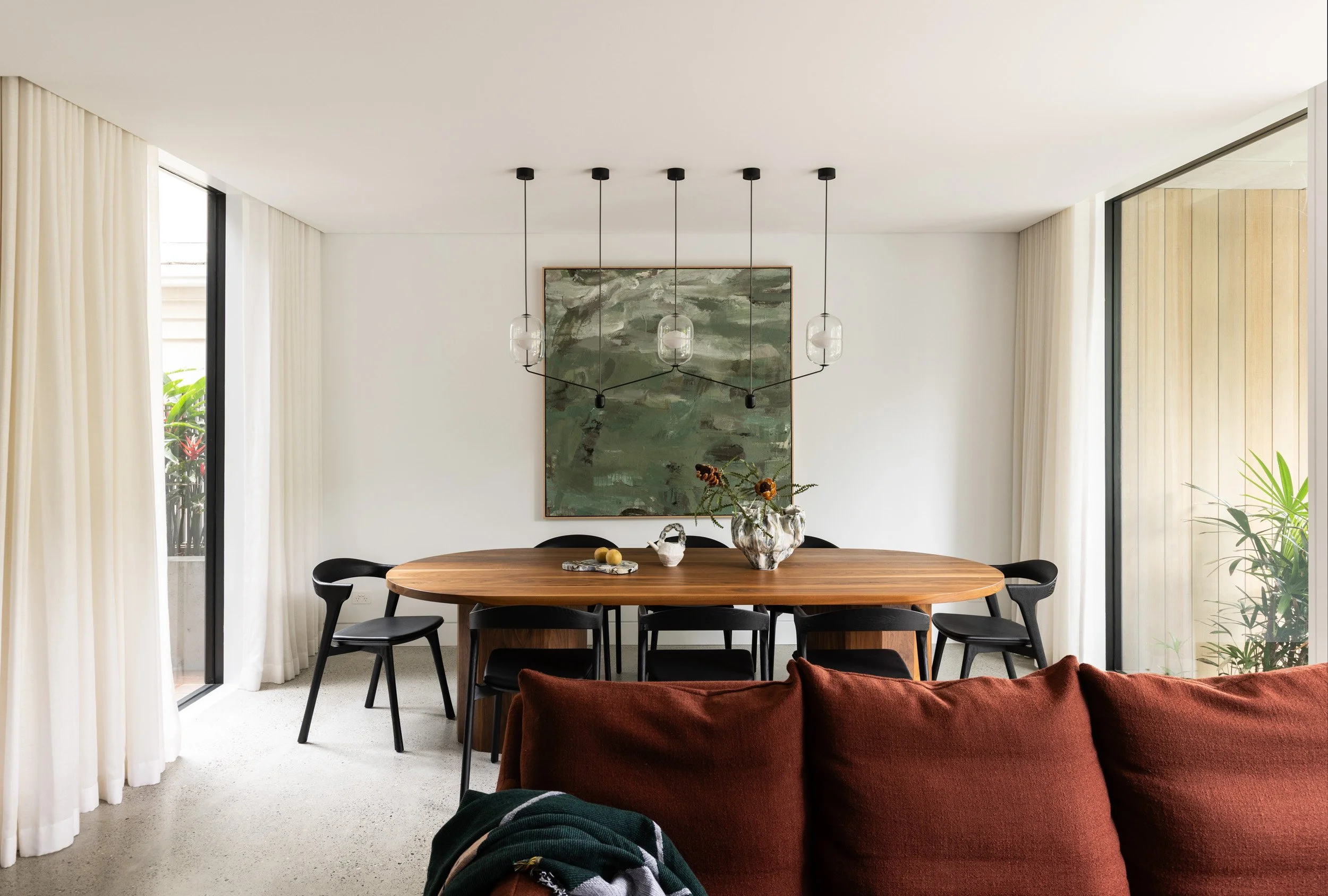 A dining room featuring a wooden table surrounded by black chairs, a large painting on the wall, and a modern chandelier with five glass pendant lights hanging from the ceiling. There are large sliding glass doors with white curtains on either side of the room, allowing natural light to fill the space, and plants visible outside.