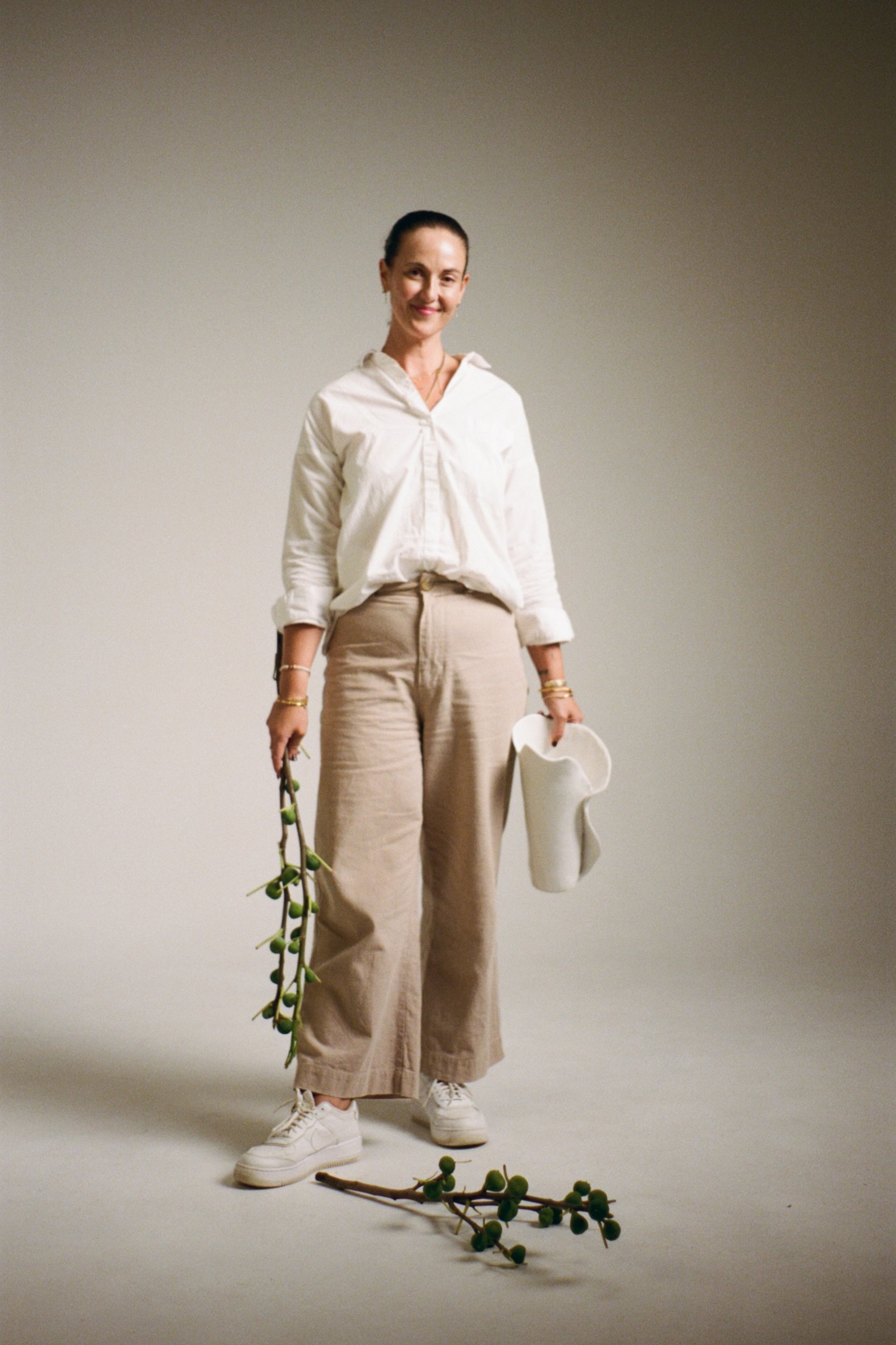 A woman with dark hair tied back, wearing a white blouse, beige wide-leg pants, and white sneakers, stands against a plain gray background. She is smiling and holding a small white bag and a branch of olive-like green fruit in her hands, with a branch on the floor in front of her.